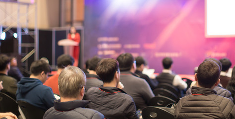Fototapete - Female Speaker Give a Speech at Corporate Conference. Youth and Senior Citizens Attend Investor and Presentation. Blurred Defocused Woman Presenter on Stage.