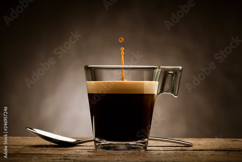 Fotografía Black coffee in glass cup with teaspoon and jumping drop, on wooden table