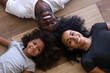 © fizkes - Happy african american family of three dad mom and little daughter portrait, smiling black child girl mother with father lying on floor looking at camera, kid parents headshot, top view from above