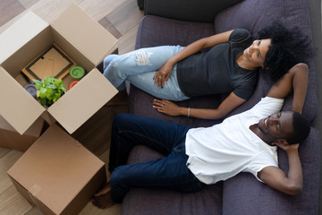 Wall Mural - African american millennial couple relaxing on couch in new home, happy young black renters tenants owners resting on sofa with boxes after relocation concept in living room, moving day, top view