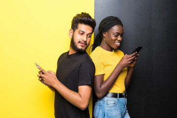 Young happy mixed race couple, indian man and african woman use phone standing back to back on the middle on different yellow and black background