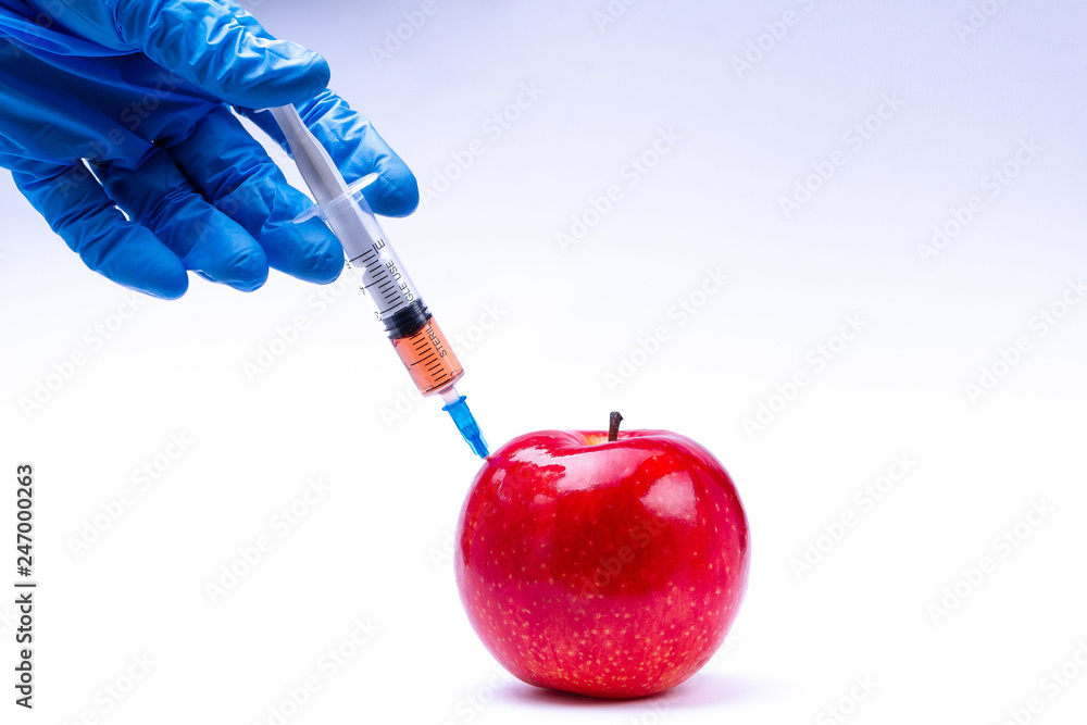 Pesticides and nitrates are injected into a red apple with a syringe. Gmo concept and genetically modified organism. Gmo free and natural fruits without chemical additives. - obrazy, fototapety, plakaty 