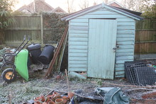 Winter Landscape Of Wooden Decaying Old Shed In Allotment Garden With Raised Beds, Wheelbarrow, Gravel Paths, Plants, Leaves, Tools, Compost Bins On Freezing Icy Day, White Frost Layer On Ground 