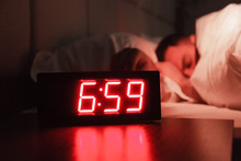 Rectangular Alarm Clock On The Bedside Table With Red Numbers, Sleeping Man In Bed In Dark Room. Concept Chef Sleeping After Hard Day At Restaurant, Sleep Disturbance, Oversleep Work, Night Shift
