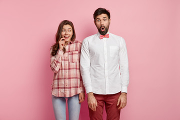 Funny two friends come on party, surprised to notice many unknown people. Smiling beautiful woman in jeans and checkered shirt stands near overwhelmed bearded guy, isolated over pink background