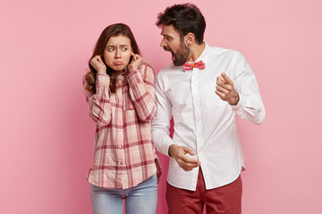 Outraged young man shouts at guilty woman and reproaches about wrong doing, blames, dissatisfied woman plugs ears, ignores loud yelling of angry husband or boss, isolated over pink background.