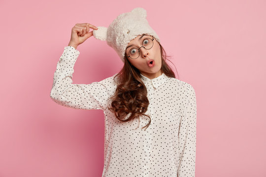 Funny surprised European young woman keeps hand on ear of hat, dressed in polka dot blouse, has folded lips, dark curly hair, isolated over pink background. People, reaction and foolishing concept