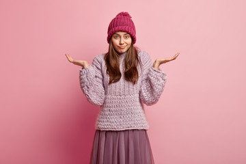 Wall Mural - Uncertain young woman with hesitant look, spreads hands, dressed in warm knitted hat, jumper and stylish shirt, isolated over pink background, expresses apathy. People, perception and attitude