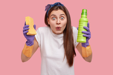 Positive young female with widely opened eyes, holds sponge and cleaning agent, dressed in casual clothes, does housework, isolated over pink background, feels surprised. Domestic work concept