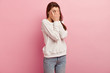 Sideways shot of surprised woman covers face with palms, peeks through fingers, has widely opened eyes, dressed in sweater and jeans, isolated over pink wall with free space for your promotion