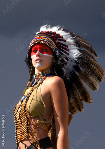 Fototapeta na drzwi i meble Młoda dziewczyna gra rolę indiańskiej Indianki. Ubrany jak rodowity Indianin w pierzastym nakryciu głowy. Pozuje na zewnątrz w otoczeniu przyrody.