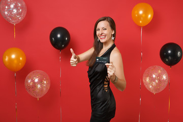 Smiling young woman in little black dress celebrating, holding credit card, showing thumb up on bright red background air balloons. Women's Day, Happy New Year, birthday mockup holiday party concept.