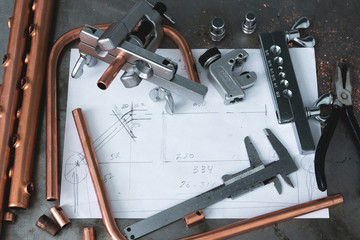 Brass pipes, pipe rammer, pipe cutter tool, calipers, pliers and a layout drafts on a fitter workbench.