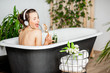 © rh2010 - Young relaxed woman listening to the music and drinking smoothie while lying in the retro bathtub at the beautiful bathroom with green plants