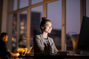 Young cheerful businesswoman sitting in dark office in front of computer monitor and networking