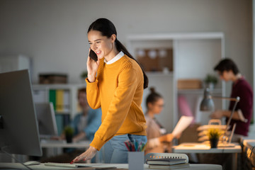 Poster - Young casual manager standing by desk in front of computer monitor, looking at its screen and talking to client on smartphone