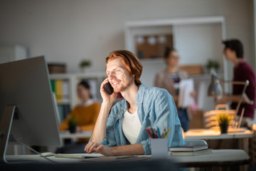 Poster - Happy young businessman with smartphone by his ear looking at computer screen while working with websites and phoning colleague