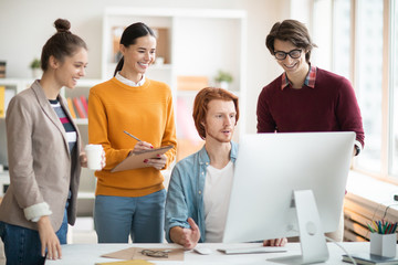 Poster - Several happy young developers standing by desk in front of computer monitor and making notes while searching on websites