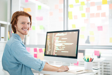 Wall Mural - young cheerful it-developer with toothy smile looking at you while sitting by workplace in front of 