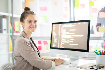 Poster - Young casual woman sitting by desk in front of camera and analyzing coded data on computer screen
