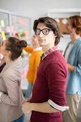 Poster - Young man in casualwear and eyeglasses standing in front of camera in working environment in office