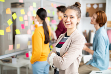 Poster - Young laughing businesswoman with her arms crossed on chest standing in front of camera in office environment