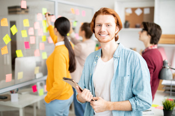 Poster - Happy young smiling businessman with notepad standing in front of camera in working environment