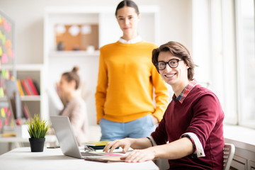 Poster - Happy casual businessman in eyeglasses sitting by desk during work on background of his colleagues