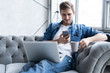 © opolja - Young man using his smartphone for online banking - sitting on sofa with laptop on leap.