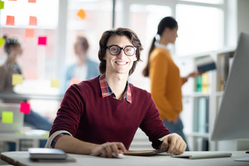 Poster - Young cheerful businessman in casualwear looking at you by desk while browsing in the net in office environment