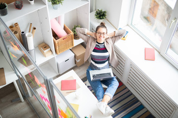 Poster - Happy young relaxed businesswoman in casualwear sitting on chair by workplace with laptop on her knees and getting inspired