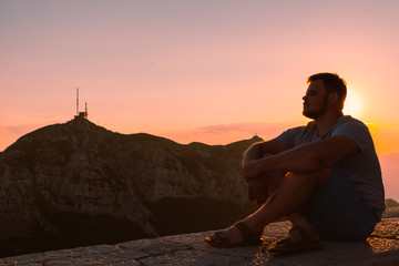 Wall Mural - young adult man sitting on stone wall with beautiful view of sunset in mountains