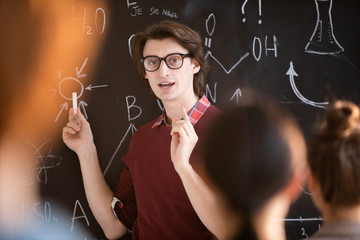 Poster - Young confident teacher in eyeglasses standing by blackboard with formula and explaining them to his students
