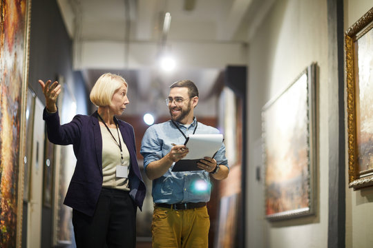 portrait of two cheerful museum workers discussing paintings walking in art gallery, copy space