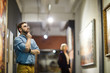 © Seventyfour - Portrait of pensive bearded man looking at paintings standing in art gallery or museum, copy space