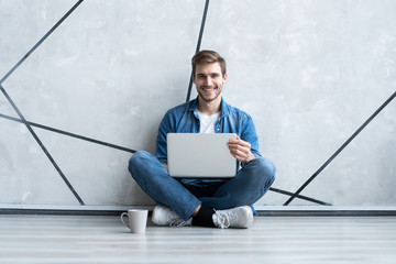 Wall Mural - Young man working on laptop sitting on floor. Guy with computer reading news, surfing net or freelancing.