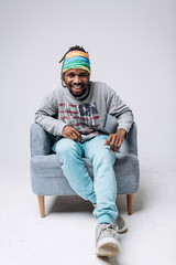 Wall Mural - Portrait of a smiling black young model in a gray sweater and blue pants on a gray background near the chair