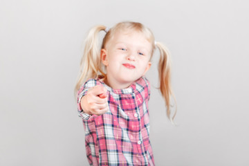 Wall Mural - Cute adorable white blonde Caucasian preschool girl making faces in front of camera. Child showing fig sign and posing in studio on plain light background. Kid expressing negative emotions