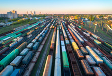 Aerial View Of Colorful Freight Trains On Railway Station At Sunset. Wagons With Goods On Railroad. Heavy Industry. Industrial Scene With Cargo Trains, City Buildings And Blue Sky. Top View From Drone