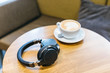 Photo of black wireless headphones and white cup of coffee on wooden table/ enjoying coffee in a cafe, music time, selective focus on headset, top view with copy space/ music and lifestyle concept.