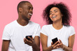 © fizkes - Happy young african american couple laughing using smartphones apps isolated on pink studio background, cheerful black friends users holding mobile phones gadgets having fun with cellphones together