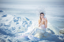 Young Model In Luxurious Strapless Corset Ball Gown Sitting On Slabs Of Broken Ice At The Frosty Seaside. Winter Fairytale Concept.