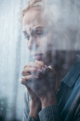 Wall Mural - sad adult woman with folded hands at home through window with raindrops