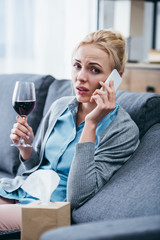 Wall Mural - woman sitting on couch, looking at camera, holding glass of red wine and talking on smartphone at home