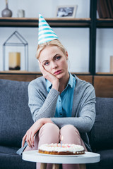 Wall Mural - sad woman in party hat propping head with hand and looking at camera while celebrating birthday at home alone