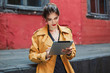 © Anton - Young attractive woman in orange trench coat and black polka dot dress thoughtfully using tablet while spending time in old courtyard