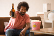© Asier - Young african man watching a movie holding a popcorn bucket and drinking a beer.