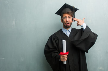 Young graduated man against a grunge wall with a copy space making a suicide gesture, feeling sad and scared forming a gun with fingers