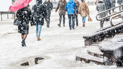 Wall Mural - Blizzard in an Urban Environment. Silhouette of a Woman with a Red Umbrella in a Crowd of Rushing People in Snowfall. Abstract Blurry Winter Weather Background.