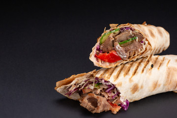 Shawarma sandwich - fresh roll of thin lavash or pita bread filled with grilled meat, mushrooms, cheese, cabbage, carrots, sauce, green. Traditional Eastern snack. On black background.